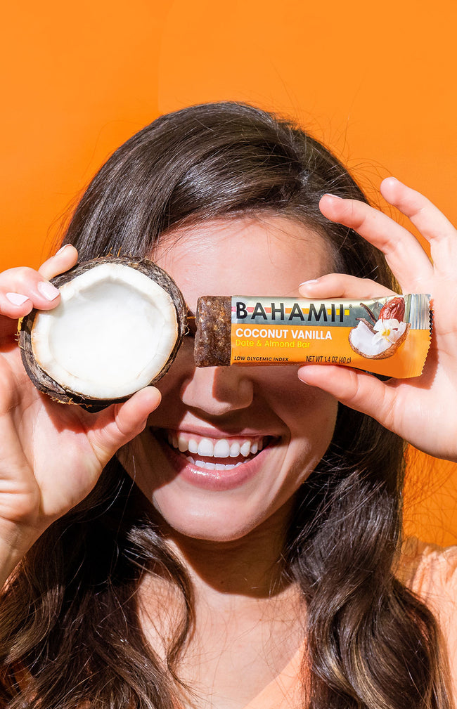 woman holding Bahami coconut vanilla date almond bar and half coconut against orange background
